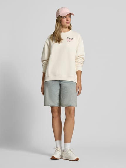 Modelka nosi bluzę Relaxed Fit Sweatshirt aus reiner Baumwolle Modell 'Classic Crew_B_4' od HUGO Blue w jasnym kremowym kolorze, luźny krój, gładka tekstura, z przodu widoczne różowe logo, do tego jasnoniebieskie szorty dżinsowe, białe sneakersy i jasnoróżowa czapka z daszkiem, Ganzkörperansicht von vorne.