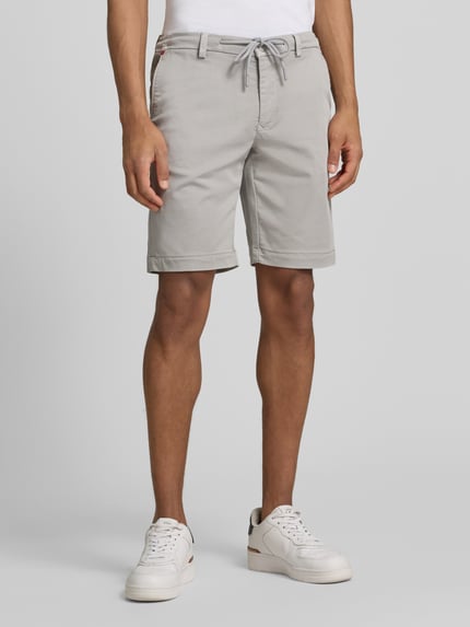 Männermodel trägt Bermudas mit Bindegürtel und französischen Eingrifftaschen von MAC, Baumwoll-Mix mit Stretch-Anteil, Straight Fit, Knopf- und Reißverschluss, Bundweite 42 cm, Innenbeinlänge 26 cm, Label-Print, helle Farbe, glatte Textur, Ganzkörperansicht von vorne.