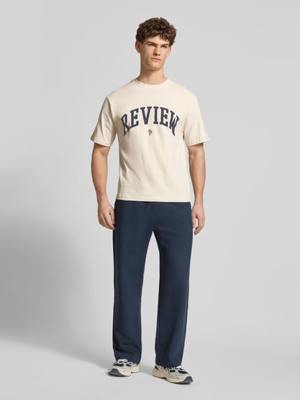 Mannelijk model draagt T-shirt met Label-Print en Rundhalsausschnitt van REVIEW, heren t-shirt van puur katoen, regular fit, labelprint, ronde hals, ruglengte bij maat M 65 cm, kleuren beige en donkerblauw, geheelbeeld van voren.