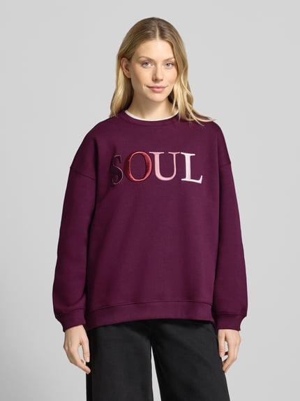 Modelka nosi Oversized Sweatshirt aus Baumwoll-Mix mit Statement-Stickerei od Smith and Soul, mieszanka bawełny, kolor śliwkowy, z napisem, okrągły dekolt, prążkowane wykończenia, bluza z długim rękawem, widok całej sylwetki z przodu.