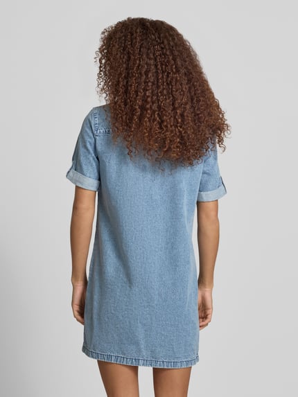 Frauenmodel trägt Jeanskleid mit Brusttaschen Modell 'NEW SIGNE' von Noisy May aus Reine Baumwolle, Gerader-Schnitt, Minikleid, Rückenlänge bei Größe S 86 cm, Brustweite 50 cm, Ärmellänge 25 cm, blauer Denim mit sichtbarer Webstruktur, Ganzkörperansicht Rückansicht.