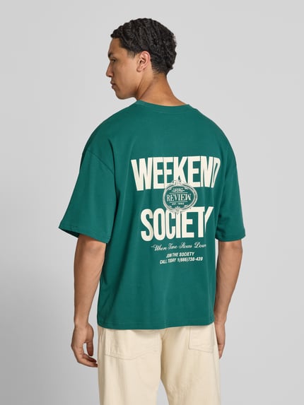 Männermodel trägt Oversized T-Shirt mit rückseitigem Print von REVIEW in Grün, Bio-Baumwoll-Mix, Oversized-Fit, Logo-Print, Ganzkörperansicht Rückansicht.