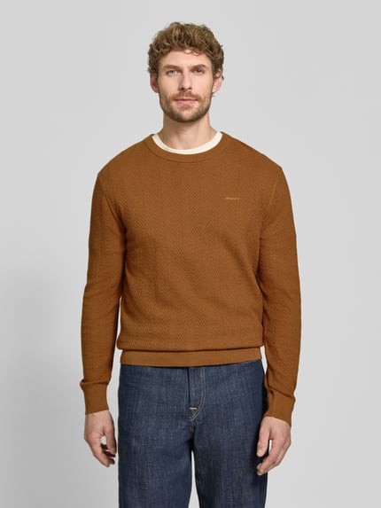 Männermodel trägt Regular Fit Pullover aus Baumwoll-Woll-Mix von GANT in Braun mit Fischgrätmuster, blaue Jeans, Ganzkörperansicht von vorne. Kontext: Herren Pullover von GANT