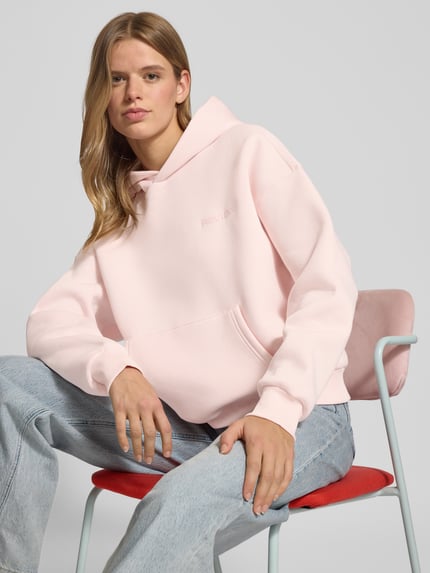 Frauenmodel trägt Oversized Hoodie mit Kapuze von Review in zartem Rosa, Baumwoll-Mix, Label-Print, Känguru-Tasche, gerippte Abschlüsse, Nahaufnahme Oberkörper.