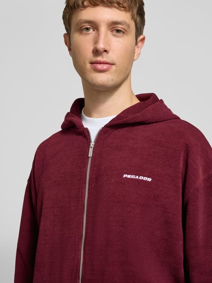 Männermodel trägt Oversized Fit Sweatjacke mit Label-Detail Modell 'Mezzano' von PEGADOR in Bordeaux, Material-Mix, Nahaufnahme Oberkörper.