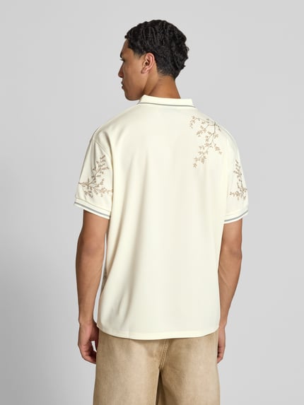 Męski model nosi koszulkę polo 'Poloshirt floralem Allover-Stitching' od Don't Waste Culture, jasny beż, krój relaxed fit, haft na całej powierzchni, nadruk z logo, kołnierzyk polo, krótki rękaw, długość tyłu w rozmiarze M: 71 cm, całkowity widok od tyłu.