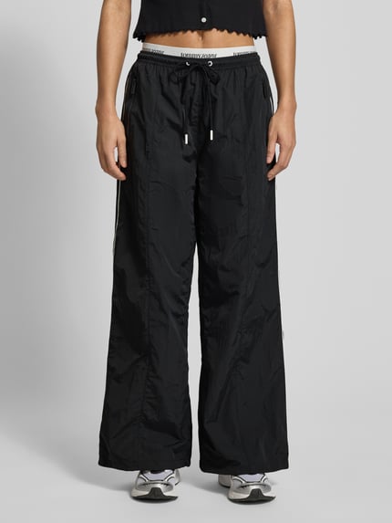 Frauenmodell trägt Regular Fit Trackpants mit Kontraststreifen von Tommy Jeans, Optik: Unifarben, elastischer Label-Bund, Tunnelzug, doppelter Bund, seitliche Reißverschlusstaschen, Gesäßtaschen, glatte Textur, Ganzkörperansicht von vorne.