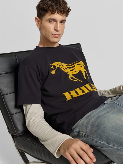Männermodel trägt T-Shirt mit Label-Print von RHUDE, reine Baumwolle, gerader Schnitt, Schwarz-Gelb, Nahaufnahme Oberkörper.
