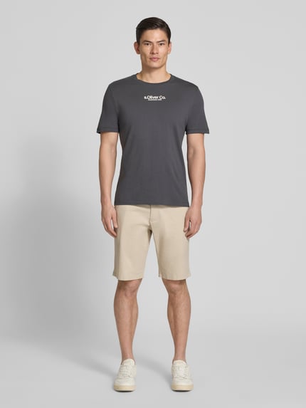 Männermodel trägt Regular Fit Bermudas aus Baumwoll-Mix Modell 'PHOENIX' von s.Oliver RED LABEL in Beige, Ganzkörperansicht von vorne, unifarben, Twill-Oberfläche, 5-Pocket-Design, cropped Länge, mit Gürtelschlaufen und Knopfverschluss.