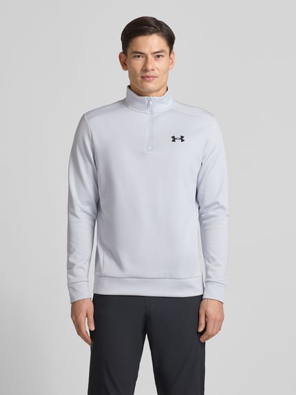 Männermodel draagt Sweatshirt met Labeldetail van Under Armour, grijs met ribboorden, los model, opstaande kraag, ruglengte maat M 72 cm, mouwlengte 67 cm, ritssluiting, labeldetail links op borst. Ganzkörperansicht van voren.