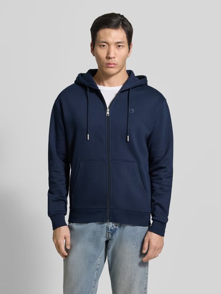 Mannelijk model draagt Regular Fit Sweatjacke aus Baumwoll-Mix van Tom Tailor Denim in effen donkerblauw, sweatmateriaal met rits, capuchon met tunnelkoord en opgestikte zakken, logo detail, geribde boorden, voorkant full-body shot.