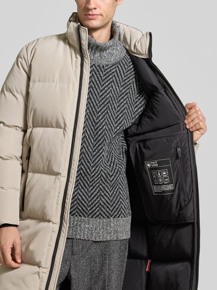 Männermodel trägt Jacke mit Kapuze und Label-Patch von NAUTGÅRD in Beige mit schwarzem Innenfutter, Nahaufnahme Oberkörper. Gerader Schnitt, Strickärmelabschlüsse, Zweiwege-Reißverschluss, Reißverschlusstaschen seitlich, Rückenlänge 100 cm, Schulterbreite 52 cm, Brustweite 60 cm, Ärmellänge 65 cm.