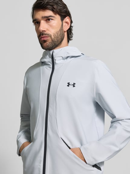Männermodel trägt Regular fit Jack met logoprint Model 'Unstoppable' van Under Armour, elastische materiaalmix, steekzakken met ritssluiting, ritssluiting, capuchon met opstaande kraag, ruglengte maat M 74 cm, mouwlengte maat M 68 cm, lichtgrijs met zwarte details, gladde textuur, nahaufnahme bovenlichaam.