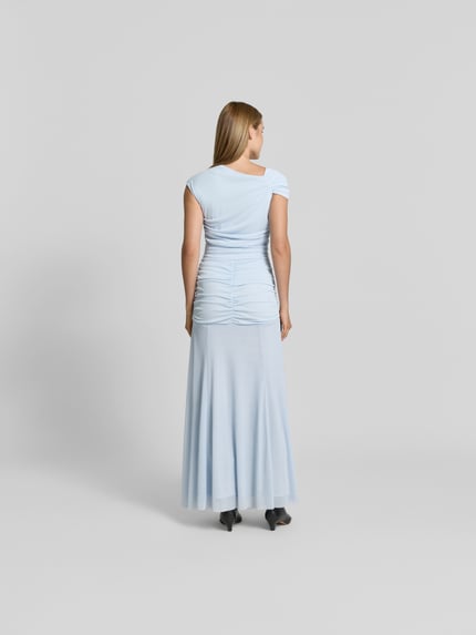 Frauenmodel trägt Maxikleid in asymmetrischem Design von ROTATE aus elastischem Material-Mix in Hellblau, Raffungen, Ganzkörperansicht Rückansicht