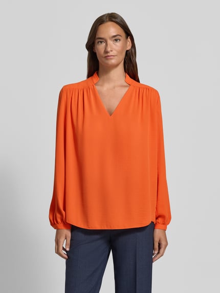 Vrouwenmodel draagt Regular Fit Bluse mit V-Ausschnitt van Marc Cain in opvallend oranje, lichte glanzende stof, lange mouwen, rechte zoom, met opstaande kraag en plooien, lengte achterzijde maat 36: 63 cm, mouwlengte maat 36: 65 cm, Ganzkörperaanzicht van voren.