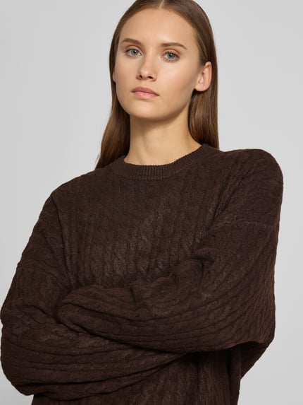 Frauenmodel trägt Oversized Fit Strickpullover mit Zopfmuster von REVIEW in Dunkelbraun, elastischer Material-Mix, Nahaufnahme Oberkörper.