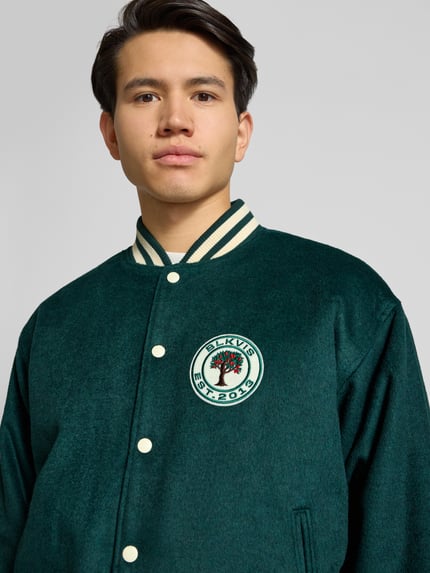Männermodel trägt College Jacke mit Label-Patch von BLKVIS in Dunkelgrün, Material-Mix mit Woll-Anteil, Nahaufnahme Oberkörper.