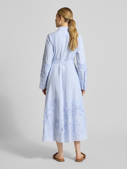 Vrouwenmodel draagt Hemdblusenkleid mit verdeckter Knopfleiste und Bindegürtel van Marc Cain, katoenmix met stretch, getailleerd model, midi-jurk, overhemdkraag, ruglengte maat 36: 130 cm, borstwijdte maat 36: 54 cm, mouwlengte maat 36: 60 cm, lichtblauwe tinten met geborduurde details, rechte vormen en gladde textuur. Ganzkörperaanzicht achteraan.
