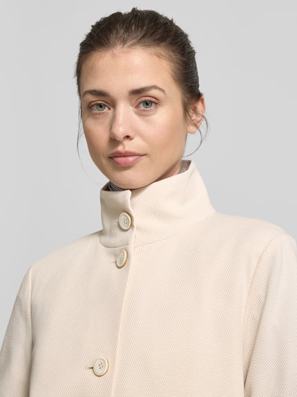 Vrouwenmodel draagt Regular Fit Jacke mit Stehkragen van WHITE LABEL in lichtbeige katoenmix met opstaande kraag, opgestikte zakken en knoopsluiting, regular fit, ruglengte maat 36: 57 cm, borstwijdte: 50 cm, mouwlengte: 65 cm, detailopname bovenlichaam.