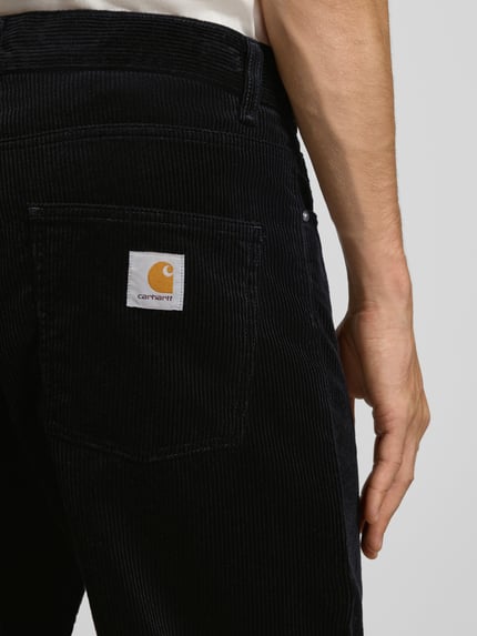 Männermodel trägt Straight Fit Hose aus reiner Baumwolle in Cord-Optik von Carhartt Work In Progress, Nahaufnahme Rückansicht. 5-Pocket-Design, schwarze Rippstruktur, sichtbares Label.