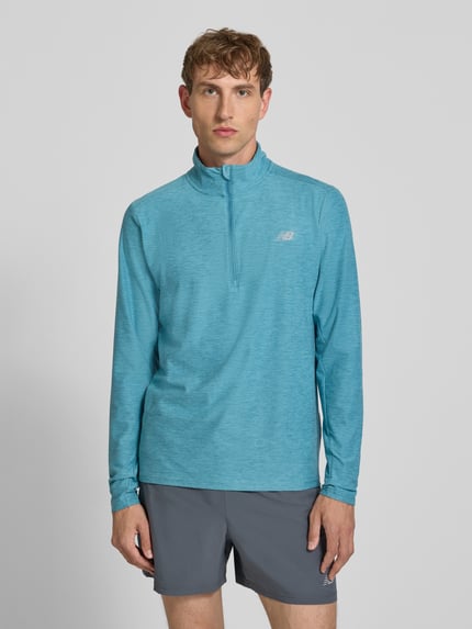 Męski model nosi Regular Fit Sweatshirt mit Troyer-Kragen von New Balance, elastyczna mieszanka materiałów, krój regular fit, nadruk z logo, długość tyłu w rozmiarze M: 69 cm, widok całej sylwetki z przodu.