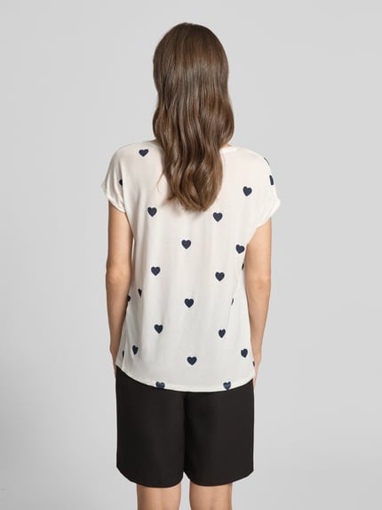 Männermodel trägt Relaxed Fit T-Shirt aus Lyocell-Mix Modell 'AVA' von Vero Moda, biały materiał z czarnym nadrukiem w serca, krótki rękaw z podwinięciem, widoczne tekstury materiału, damska koszulka z okrągłym dekoltem, allover print, widok całej sylwetki z tyłu.