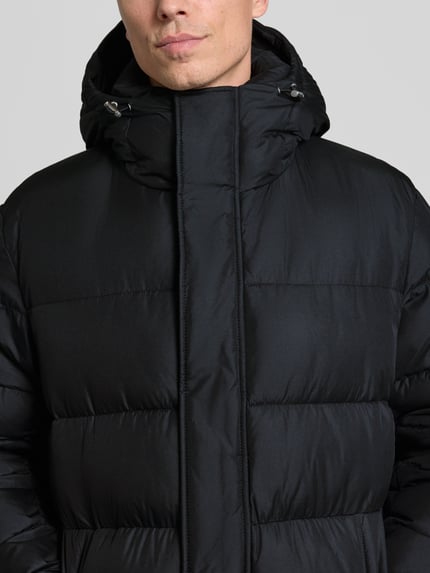 Männermodel trägt Steppmantel mit Kapuze Modell 'H-Carpa' von BOSS in Schwarz, sichtbare Nähte, verdeckter Reißverschluss, Nahaufnahme Oberkörper.