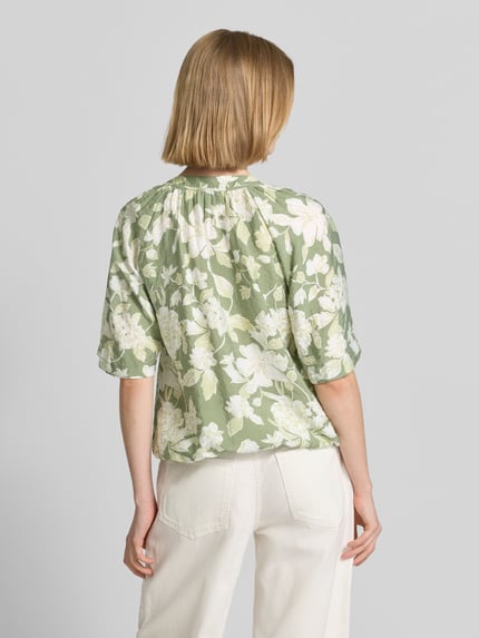 Frauenmodel trägt Relaxed Fit Blusenshirt aus Viskose-Mix von s.Oliver RED LABEL, Optik:Allover-Muster, florales Muster, Crinkle-Design, Hellgrün und Weiß, Kurzarm, Raglanärmel, Bindeband, Rumpfabschluss elastisch, Ganzkörperansicht Rückansicht.