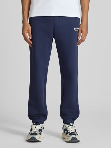 Męski model nosi spodnie dresowe Straight Fit Sweatpants aus Baumwoll-Mix Model 'Kane' von JACK & JONES, mieszanka bawełny, kieszeń z tyłu, detal z logo, elastyczny pas i zakończenia nogawek, długość wewnętrzna nogawki w rozmiarze M 72 cm, widok z przodu na całe ciało.