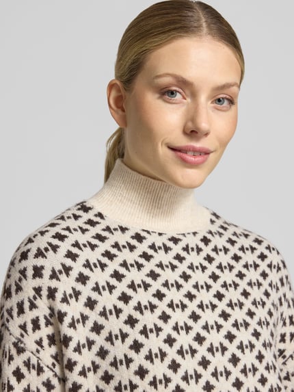 Frauenmodel trägt Pullover mit Jacquardstruktur Modell 'VMBIRCH' von VERO MODA in Creme mit braunem grafischen Allover-Muster, Stricktextur, hochgeschlossenem Mock Neck, Nahaufnahme Oberkörper.