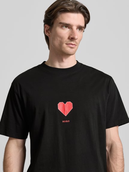 Männermodel trägt Regular Fit T-Shirt aus reiner Baumwolle Modell 'DALENTIN' von HUGO in Schwarz mit rotem Herzprint, Nahaufnahme Oberkörper.