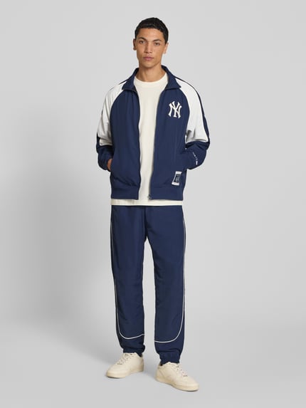 Männermodel trägt Relaxed Fit Tracksuit mit Logo-Patch von Champion, blau mit weißen Details, elastische Abschlüsse, Ganzkörperansicht von vorne.