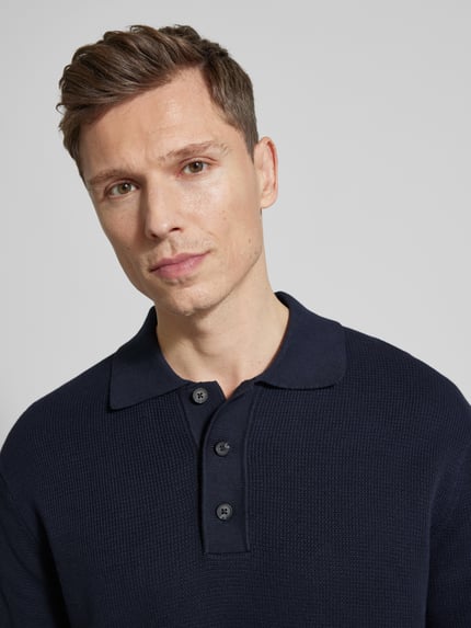 Männermodel trägt Relaxed Fit Poloshirt aus reiner katoen Modell 'DANE' van SELECTED HOMME in effen, gebreide look met v-hals en polokraag, label stitching, korte knoopsluiting, korte mouwen met geribde boorden, detailopname bovenlichaam.