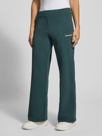 Frauenmodel trägt Flared Fit Jogginghose aus mieszanka bawełny Model 'Anni' von PEGADOR, elastyczny pas, wpuszczane kieszenie, wyhaftowane logo, Długość wewnętrzna nogawki w rozmiarze S: 82 cm, zielony, gładka faktura, Ganzkörperansicht von vorne.