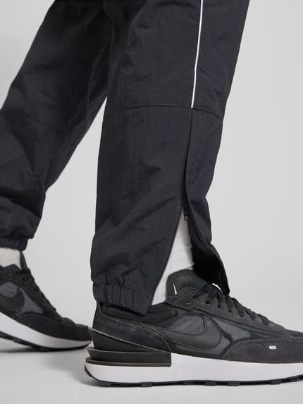 Männermodel trägt Trainingshose mit Logo und elastischem Bund von Nike in Schwarz, Relaxed-Fit, elastische Beinabschlüsse, Nahaufnahme Bein, Innenbeinlänge bei Größe M: 76 cm.