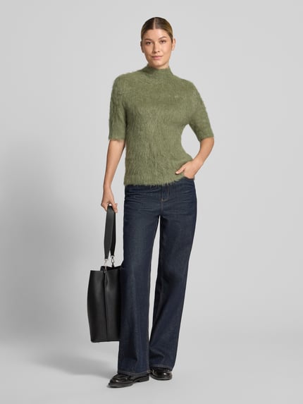 Frauenmodel trägt Strickshirt aus Alpaka-Mix mit kurzem Stehkragen von someday, körpernaher Schnitt, grüner Farbton, weiche Textur, kombiniert mit dunkler Hose und schwarzer Tasche, Ganzkörperansicht von vorne.