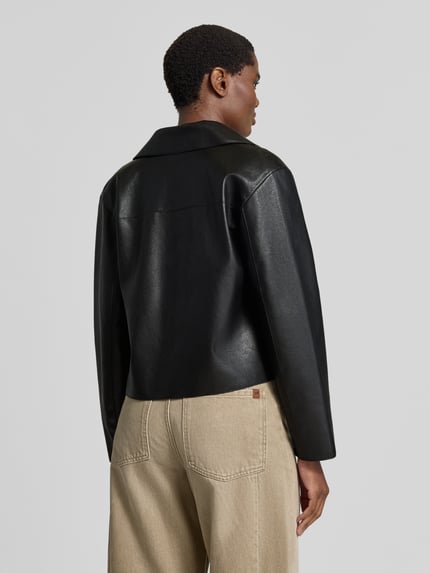 Frauenmodel trägt Jacke in Leder-Optik Modell 'SABINA' von MANGO, schwarze Oberfläche, beige Hose, Rückansicht, Ganzkörperansicht.
