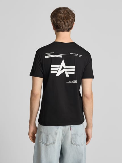 Männermodel trägt T-Shirt mit Label-Patch von Alpha Industries in Schwarz, Reine Baumwolle, Gerader-Schnitt, Rückenlänge 66 cm, Ganzkörperansicht Rückansicht.