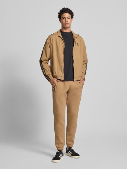 Męski model nosi Sweatpants mit elastischem Bund od adidas Originals, luźny krój, beżowy, kieszenie, widok całej sylwetki z przodu.