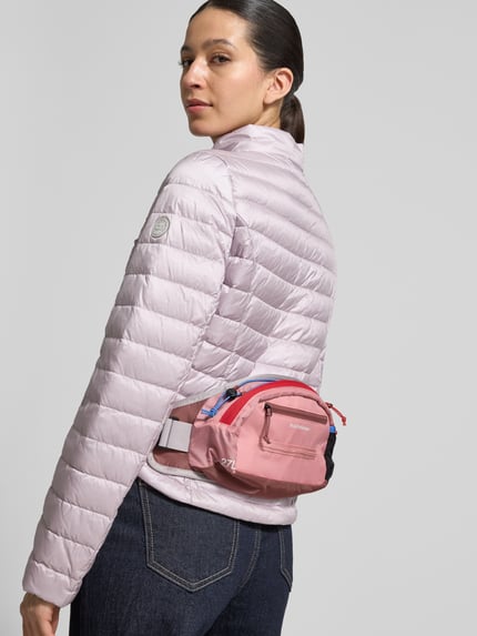 Frauenmodel trägt Bauchtasche mit Label-Detail von PEAK PERFORMANCE in Rosa mit Reißverschluss, Nebenfächer, verstellbarem Riemen, Ganzkörperansicht Rückansicht. Breite 41 cm, Länge 18 cm, Tiefe 7 cm, Riemenlänge 65 cm, Innenfächer.