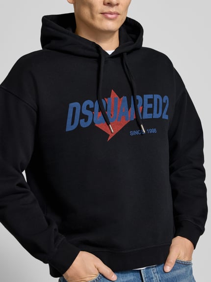 Männermodel trägt Relaxed Fit Hoodie aus reiner Baumwolle von Dsquared2 in Schwarz mit blau-rotem Label-Detail, Nahaufnahme Oberkörper.
