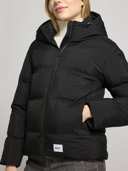 Vrouwenmodel draagt zwarte gewatteerd jack met capuchon, tunnelkoord, ritssluiting, labeldetail, Model 'VEGAS' van khujo, regular fit, nahaufnahme bovenlichaam. Ruglengte (S): 61 cm, schouderbreedte (S): 49 cm, borstwijdte (S): 49 cm, mouwlengte (S): 55 cm.
