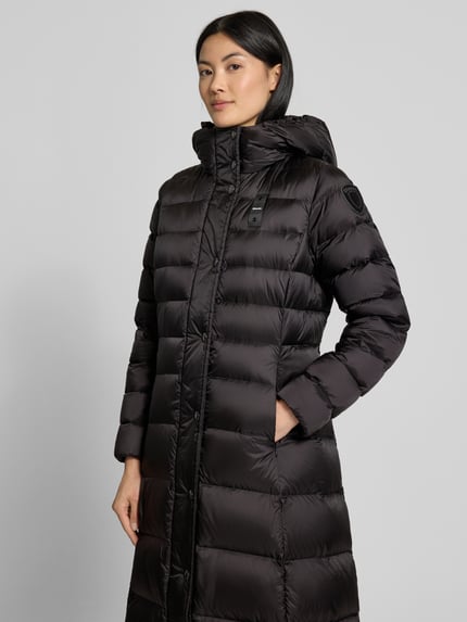 Kobieta modelka nosi płaszcz puchowy z efektem pikowania od Blauer USA, model 'Anita', detal z logo, kaptur ze stójką, wpuszczane kieszenie z zamkiem, czarny, taliowany krój, długość tyłu w rozmiarze S: 114 cm, szerokość ramion w rozmiarze S: 36 cm, szerokość w klatce piersiowej w rozmiarze S: 49 cm, długość rękawa w rozmiarze S: 60 cm, widok całej sylwetki z przodu.