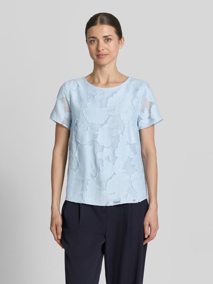 Frauenmodel trägt Blusenshirt aus Viskose-Mix mit Strukturmuster von More & More in Hellblau mit geradem Schnitt und Rundhalsausschnitt, Ganzkörperansicht von vorne.