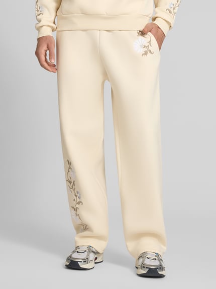 Männermodel trägt Straight Fit Sweatpants mit floraler Stickerei von REVIEW in Beige mit elastischem Bund, Ganzkörperansicht von vorne.