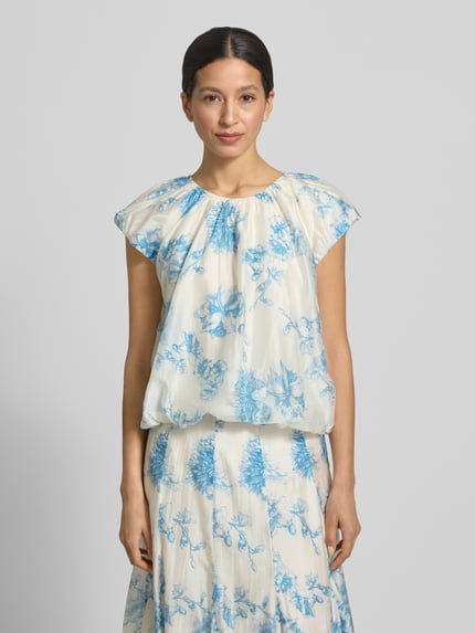 Frauenmodel trägt Regular Fit Bluse aus Lyocell-Mix Modell 'Evangelia' von MSCH Copenhagen, floraler Allover-Print, Falten-Optik, hellblau auf cremeweiß, kurzärmelig, Rückenlänge bei Größe S 50 cm, Ganzkörperansicht von vorne.