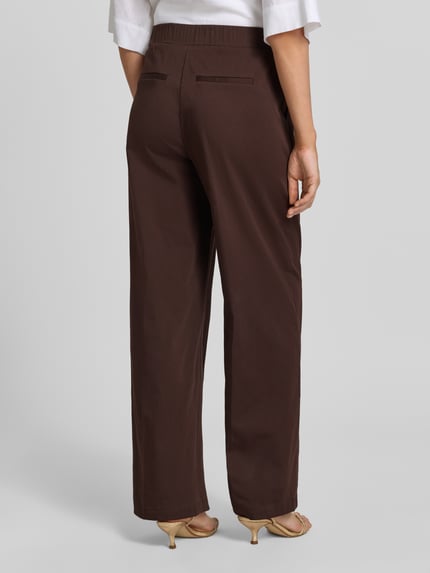 Männermodel trägt spodnie materiałowe z mieszanki bawełny z dodatkiem streczu, krój loose fit, francuskie wpuszczane kieszenie, model 'Mucia Base' od OPUS PANTS, kolor brązowy, elastyczny pas, tył kieszenie z wypustką, długość wewnętrzna nogawki 80 cm w rozmiarze 36, szerokość w rozmiarze 36: 31 cm, zdjęcie całej sylwetki z tyłu.