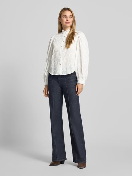 Frauenmodel trägt Bluse aus reiner Baumwolle mit Stehkragen Modell 'ROMA' von MANGO, weiße Stickerei, ballonartige Ärmel, gerade Passform, Ganzkörperansicht von vorne.