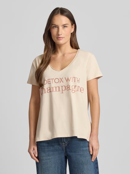 Frauenmodel trägt Loose Fit T-Shirt mit V-Ausschnitt von miss goodlife, beigefarbener Baumwoll-Elasthan-Mix, Statement-Print, Nahaufnahme Oberkörper.
