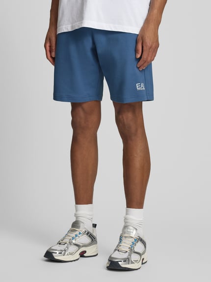 Männermodel trägt Sweatshorts mit Logo und Eingrifftaschen von EA7 Emporio Armani in Blau, Reine Baumwolle, Regular-Fit, Nahaufnahme Beine, Ganzkörperansicht von vorne.